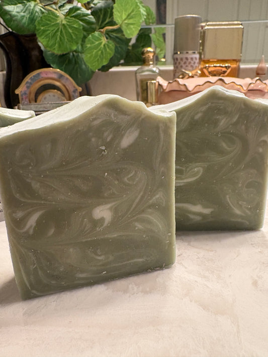 Two handmade rectangular bars of soap with a sage green and white marbled swirl pattern, displayed on a light marble countertop with a blurred background of a plant and decorative items.