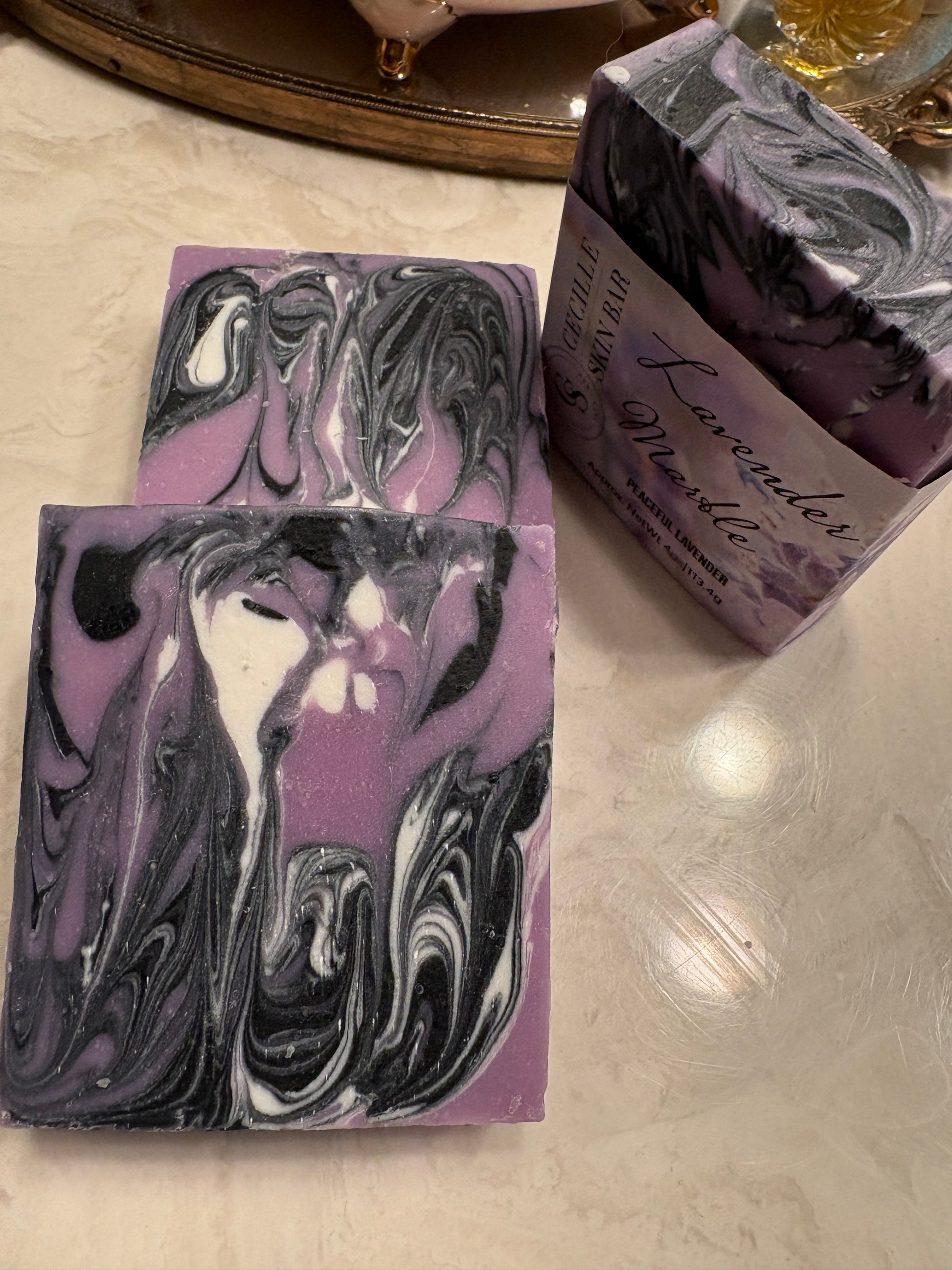 Lavender marbled soap bars and a matching wrapped bar box on a light marble surface. The soaps have a purple, black, and white swirl pattern, and the box is labeled 'Lavender Marble'.