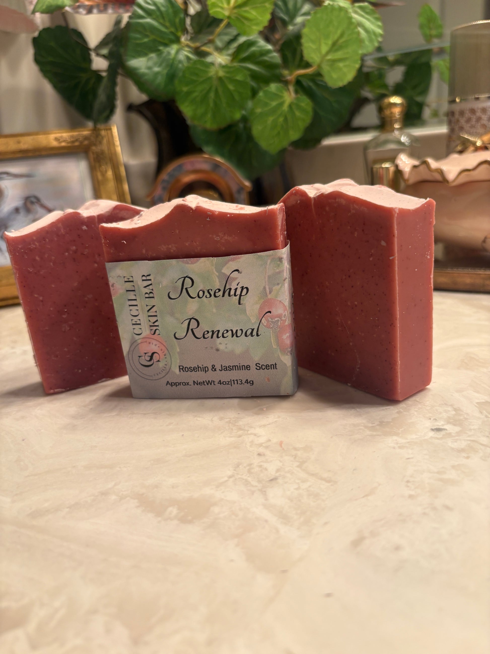 Three handmade soap bars in a dusty rose pink color, with a central bar showing a label that reads 'Rosehip Renewal' and 'Succulent Skin Bar', set on a light marble surface with a blurred background of a plant and home decor.