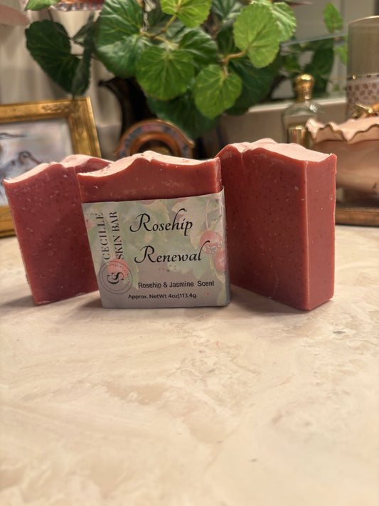 Three handmade soap bars in a dusty rose pink color, with a central bar showing a label that reads 'Rosehip Renewal' and 'Succulent Skin Bar', set on a light marble surface with a blurred background of a plant and home decor.