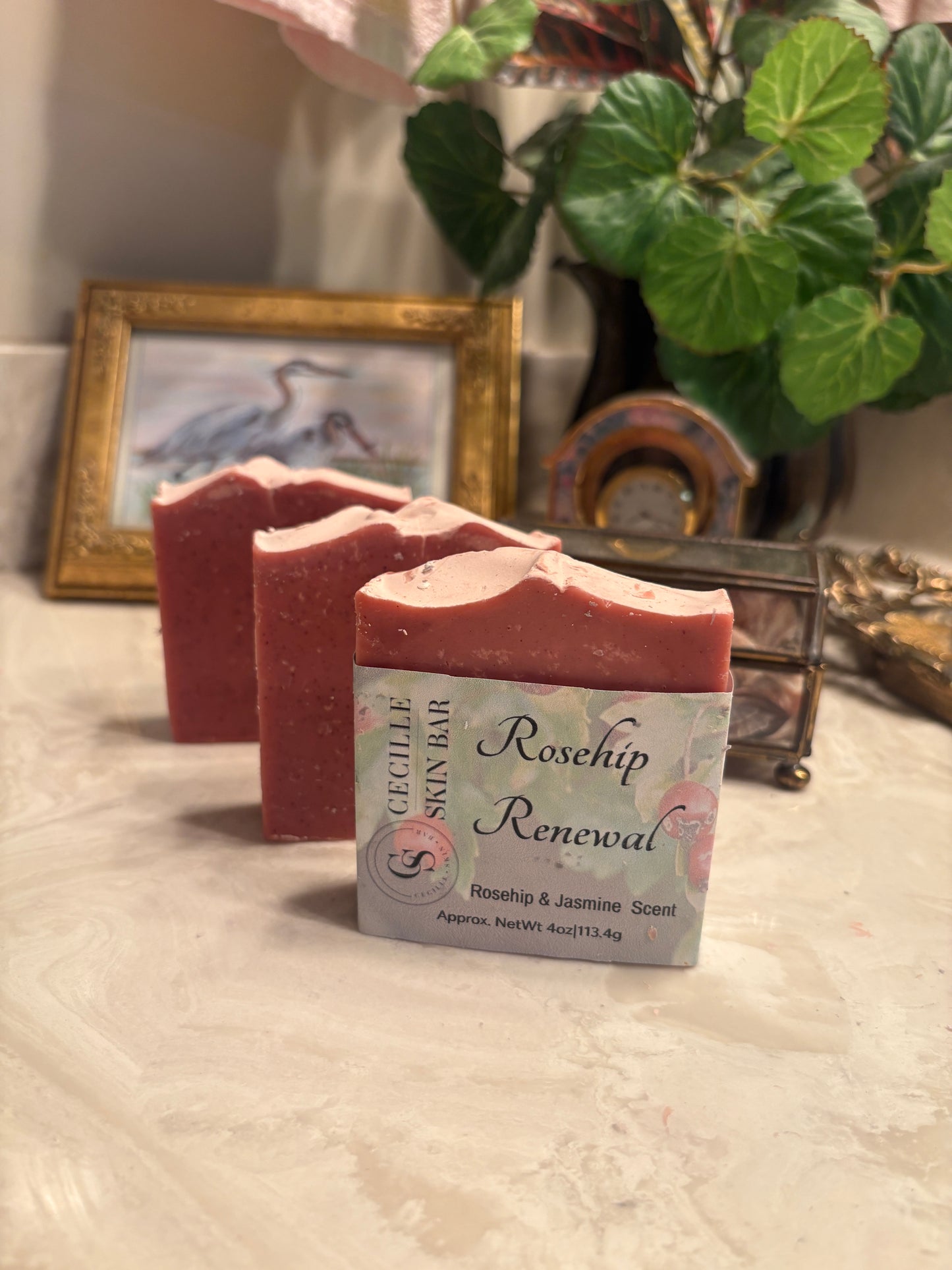 Three handmade soap bars in a dusty rose pink color, with one bar in front showing a label that reads 'Georgette Skin Bar Rosehip Renewal'. The soaps are arranged on a textured white surface with a decorative background containing a framed picture, a plant, and a small glass container.