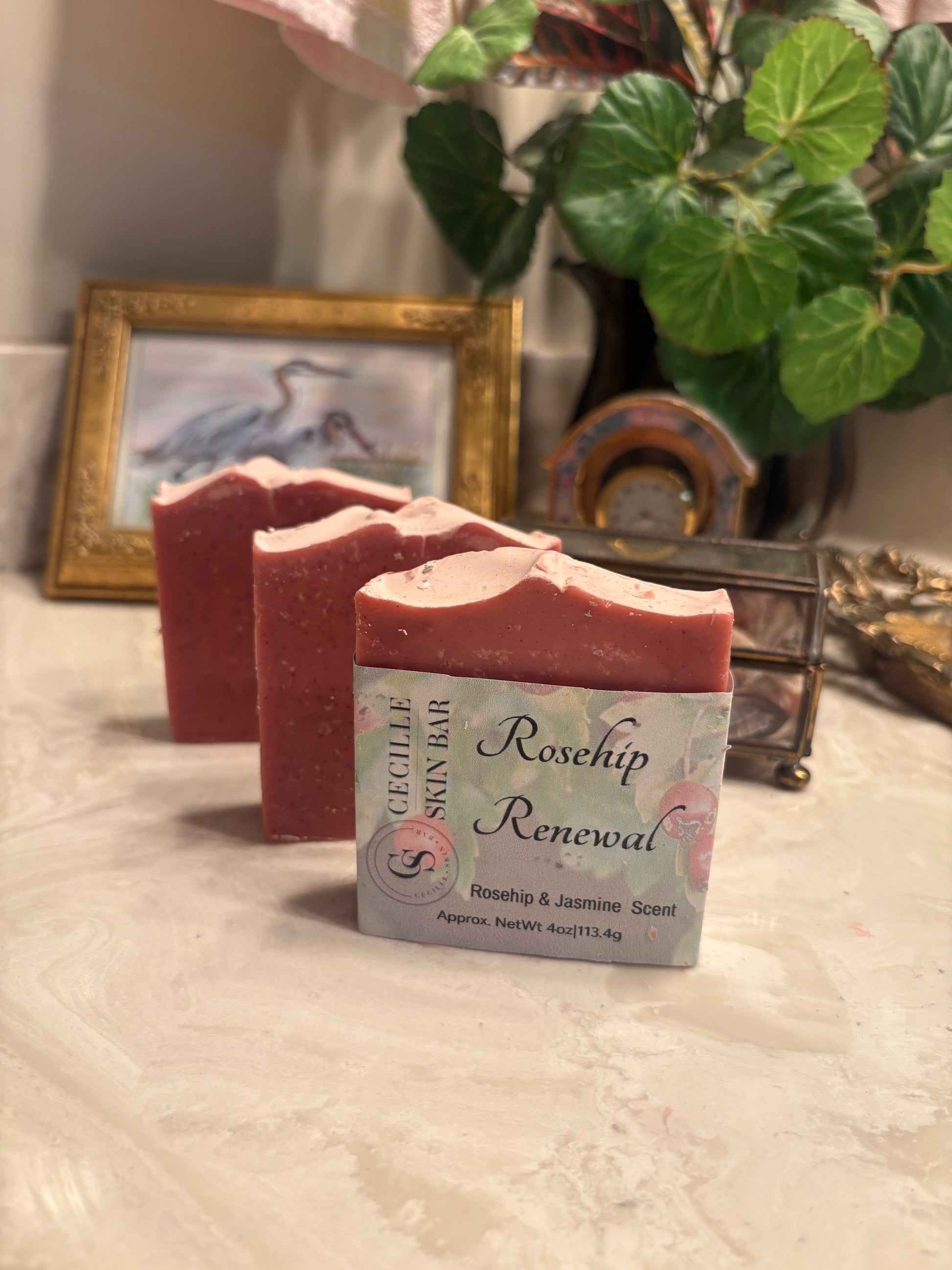 Three handmade soap bars in a dusty rose pink color, with one bar in front showing a label that reads 'Georgette Skin Bar Rosehip Renewal'. The soaps are arranged on a textured white surface with a decorative background containing a framed picture, a plant, and a small glass container.