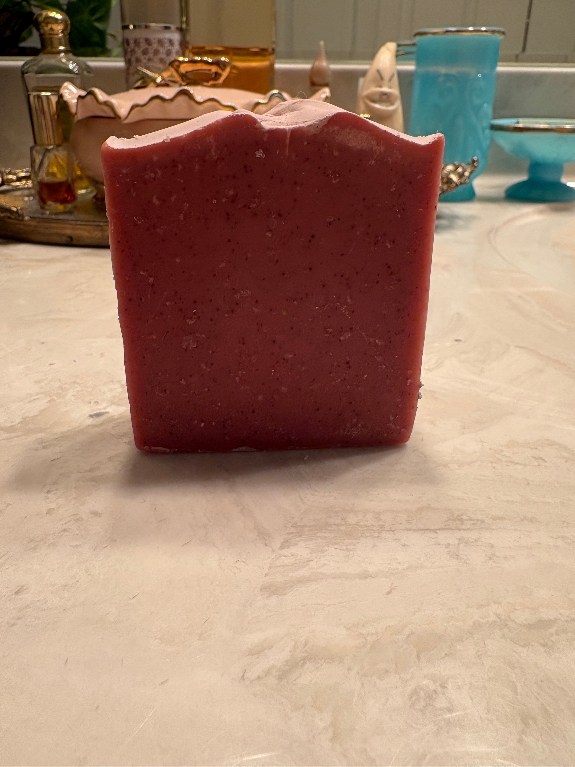 A rectangular bar of handmade soap with a scalloped top edge, standing upright on a light marble countertop. The soap has a textured, speckled surface in a deep berry pink color.