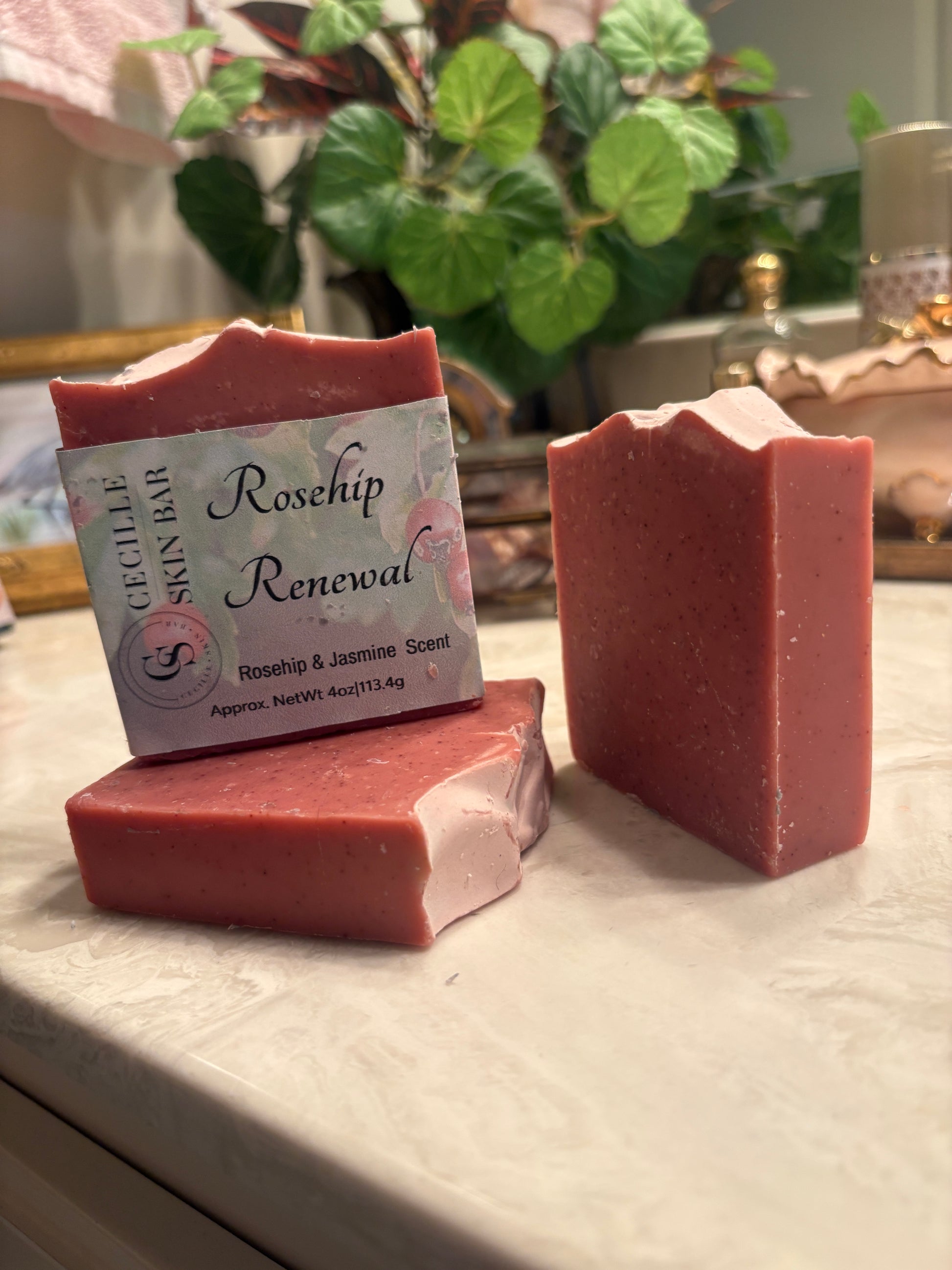 Three pink rosehip soap bars on a light marble surface, with one bar standing upright and showing a label that reads 'Cecile's Skin Bar Rosehip Renewal'.