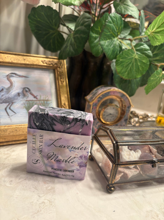 A bar of Cerelle Skin Bar Lavender Marble soap sits on a light-colored surface next to a small glass jewelry box, with a framed picture, a clock, and a potted plant in the background.