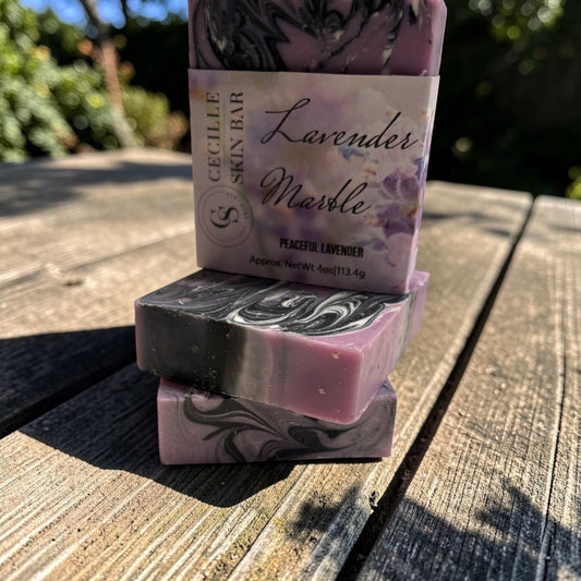 Three stacked handmade lavender soap bars on a weathered wooden table outdoors. The top bar is wrapped in a paper label reading 'Cecille Skin Bar Lavender Marble Peasicc Lavender'.