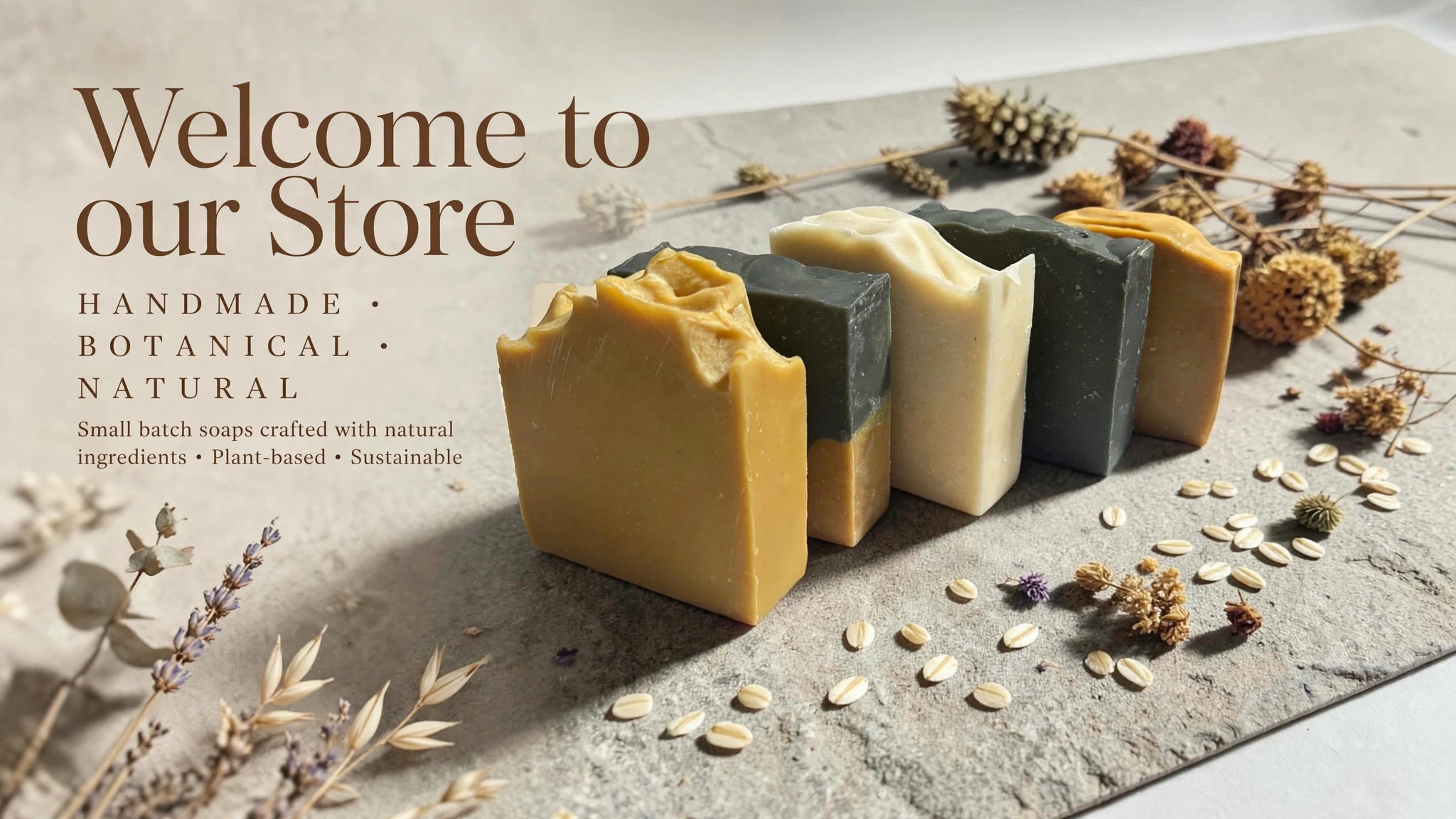 Four handmade rectangular bars of soap in yellow, cream, and dark green are arranged on a stone surface with dried botanicals and oats. Large text on the left reads 'Welcome to our Store' with smaller text describing the soaps as handmade, botanical, and natural.